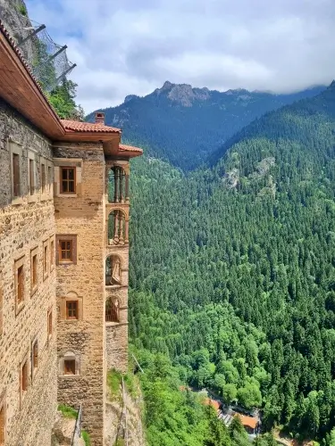Trabzon Sümela Manastırı’nın dış cephe hücreleri ve ahşap balkonları; Altındere Vadisi’nin yoğun orman dokusu ve dağ manzarasıyla birlikte yüksek kayalıktan görülen panorama.